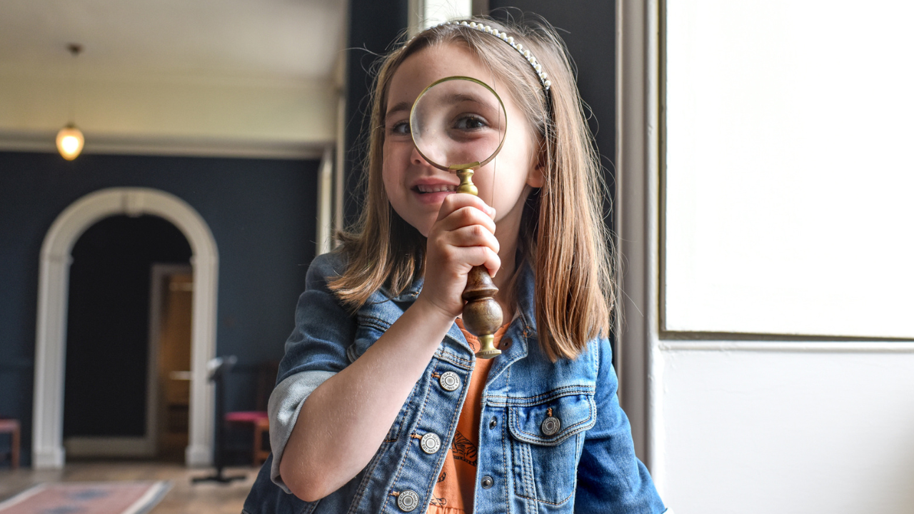 Girl holding up a magnifying glass with a long corridor behind her