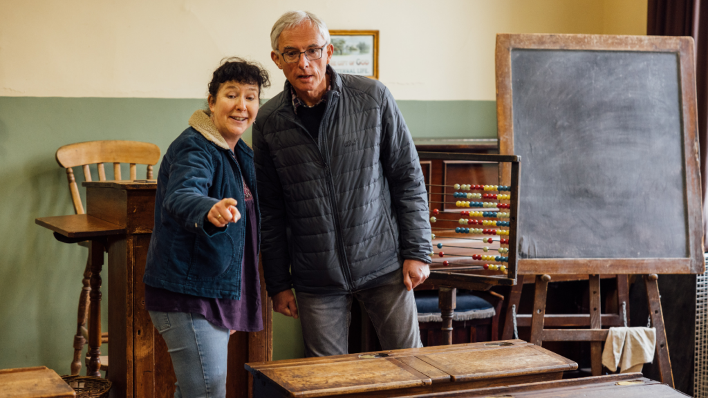 An older man and woman in a museum's Victorian schoolroom excitedly pointing to something out of shot