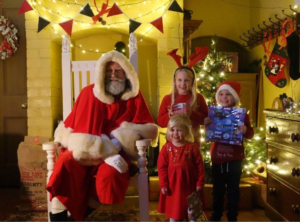 Two girls and a boy in Christmas outfits posing with Santa and their presents in a Santa's grotto