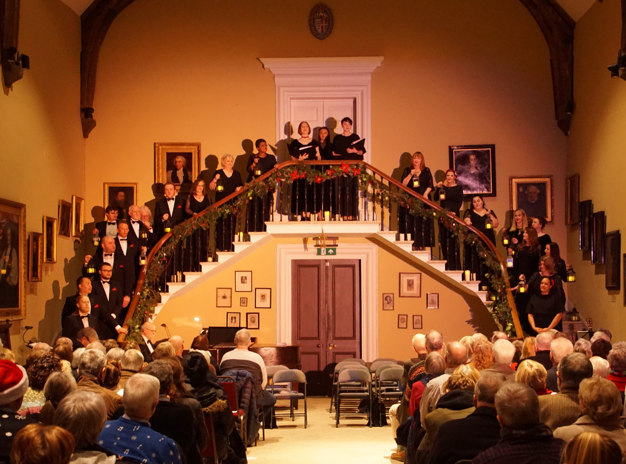 Smartly dressed people performing to an audience from the Hartlebury Castle Great Hall staircase, while holding lanterns