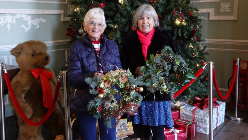 Two women in front of a Christmas tree, holding Christmas wreaths