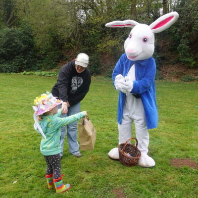 Girl wearing an Easter bonnet shows the Easter Bunny her bag of plastic eggs outside as her adult watches
