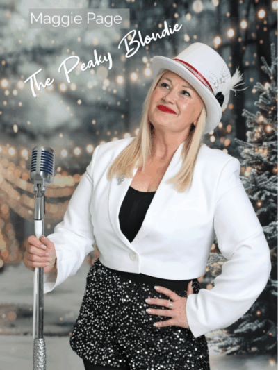 Maggie Page, The Peaky Blondie. Woman posing with a microphone and wearing a festive outfit, in front of a festive background.