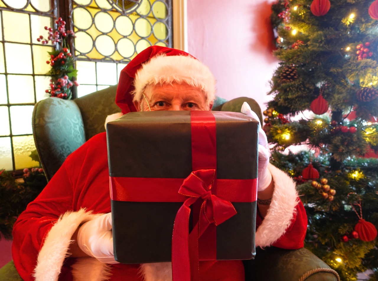 Santa hiding behind a large present, sitting in his grotto with a stained glass window and Christmas tree behind him