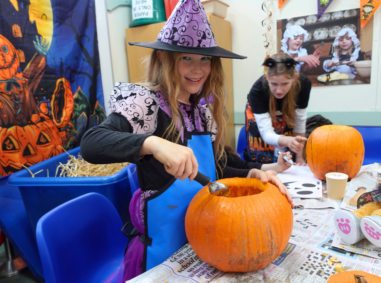 A girl in a witch's hat carves a pumpkin and smiles, while a female staff member helps a child in the background