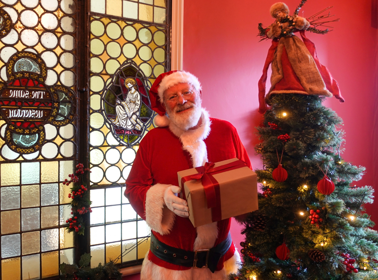 Santa in his grotto holding a present, with a stained glass window and a Christmas tree behind him