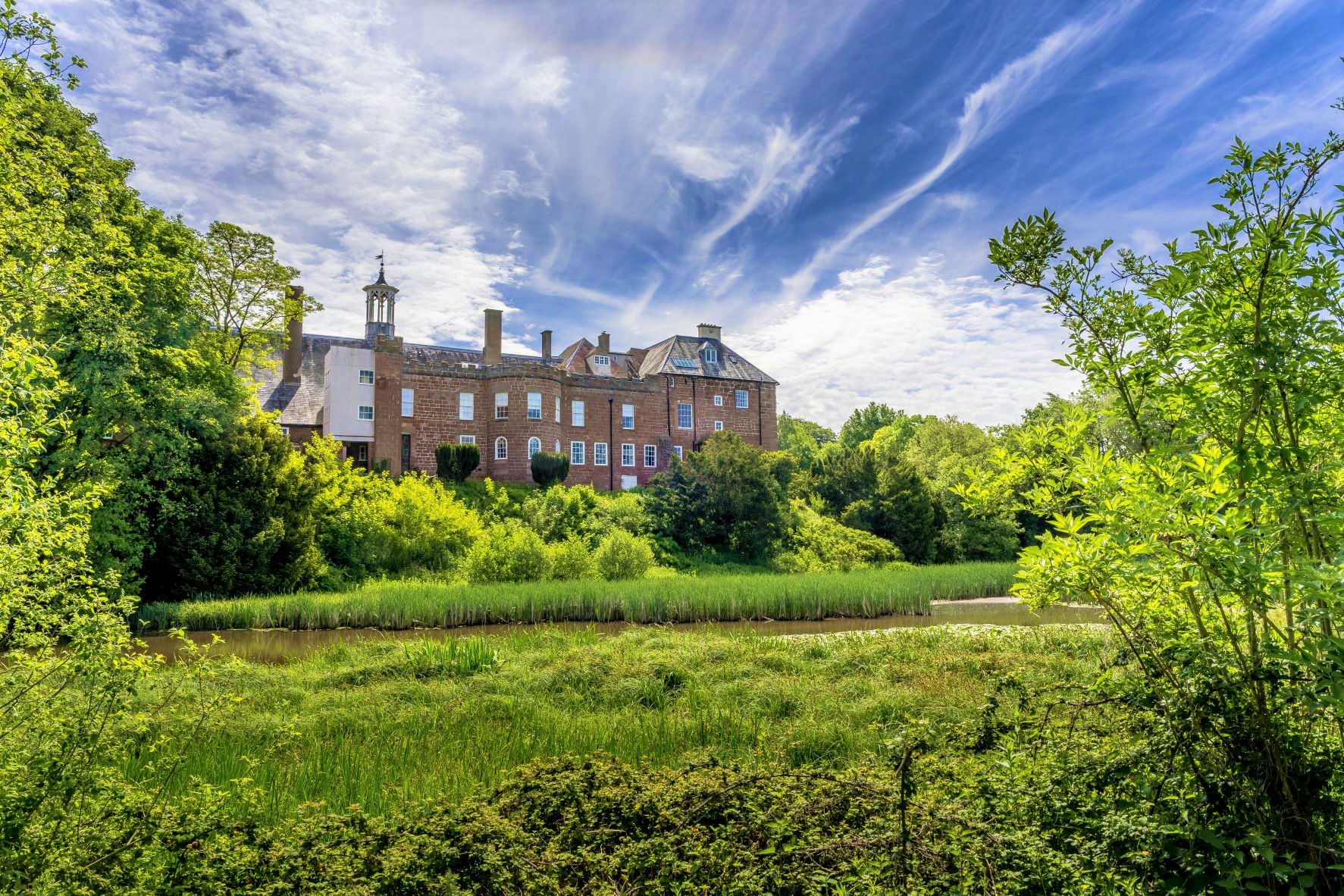 Hartlebury Castle as seen from the moat, with green foliage and trees, plus a blue and cloudy sky