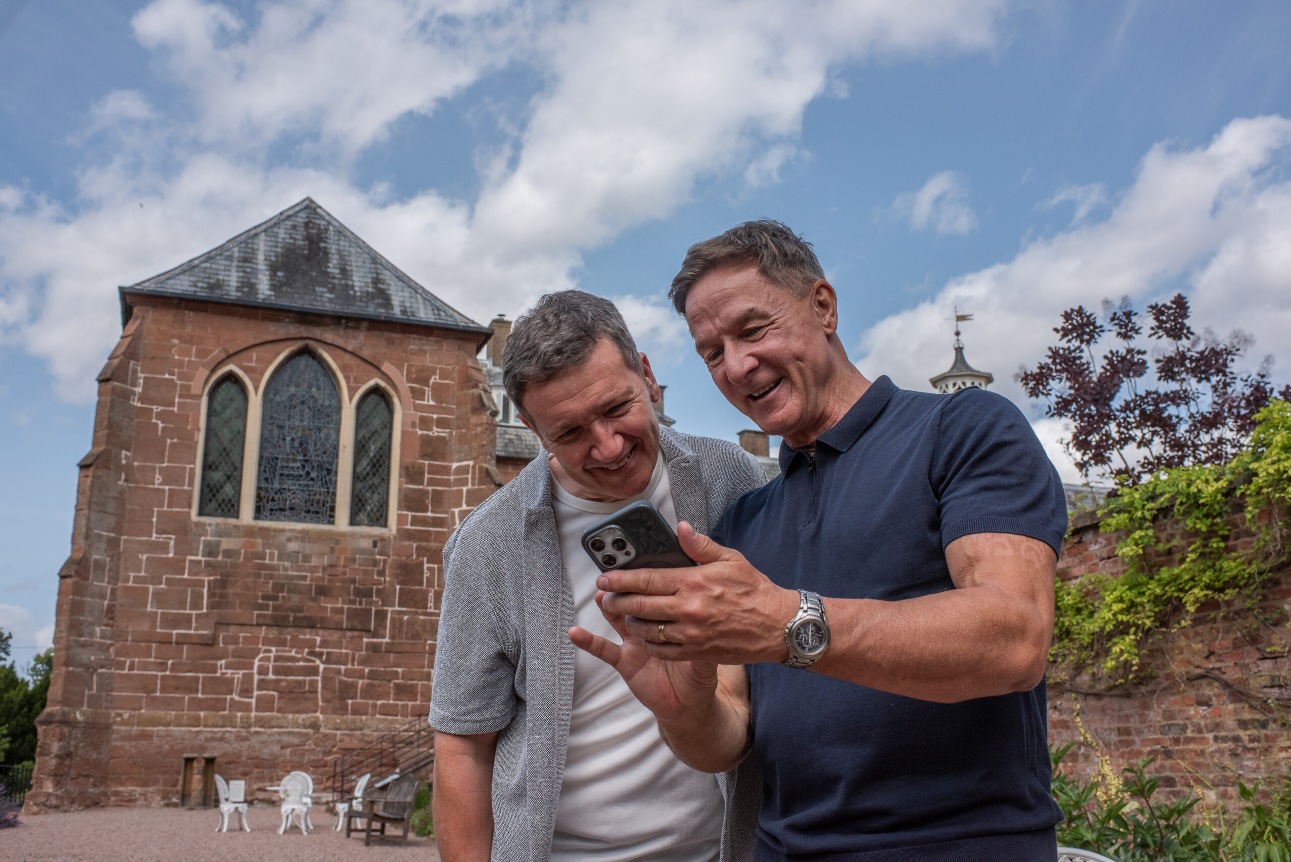 Two middle-aged men smile down at a mobile phone with the exterior of the Hartlebury Castle Chapel behind them