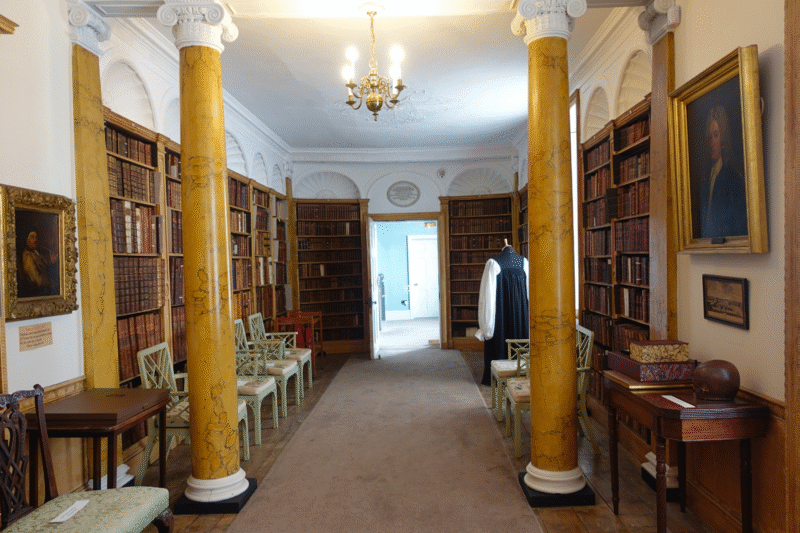 Corridor of the Hurd Library facing the glass door, and featuring bookshelves and marble effect columns