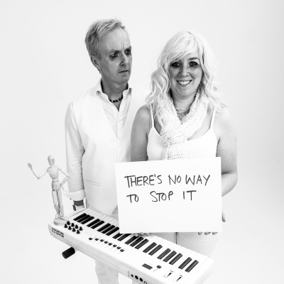 The Rude Awakening black and white promotional photo. A man and woman with heavy eye makeup, dressed in white and standing by a small keyboard, with the woman holding a sign saying 'There's no way to stop it'.