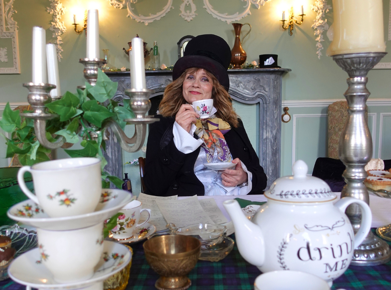 Female Mad Hatter posing with a tea cup while sat at a table set for a tea party, featuring a teapot that says 'drink me'.