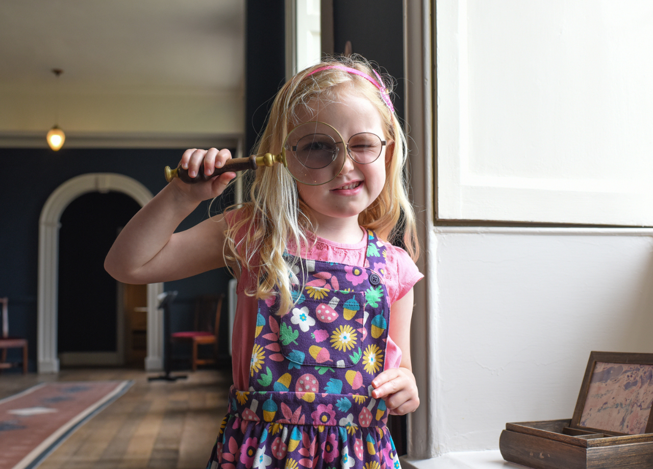 Girl looking through magnifying glass with the corridor of the Castle's Long Gallery behind her