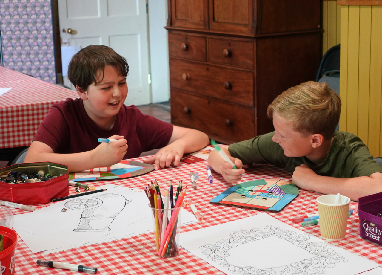 Two boys smiling at each other while colouring on wooden shields at a gingham craft table