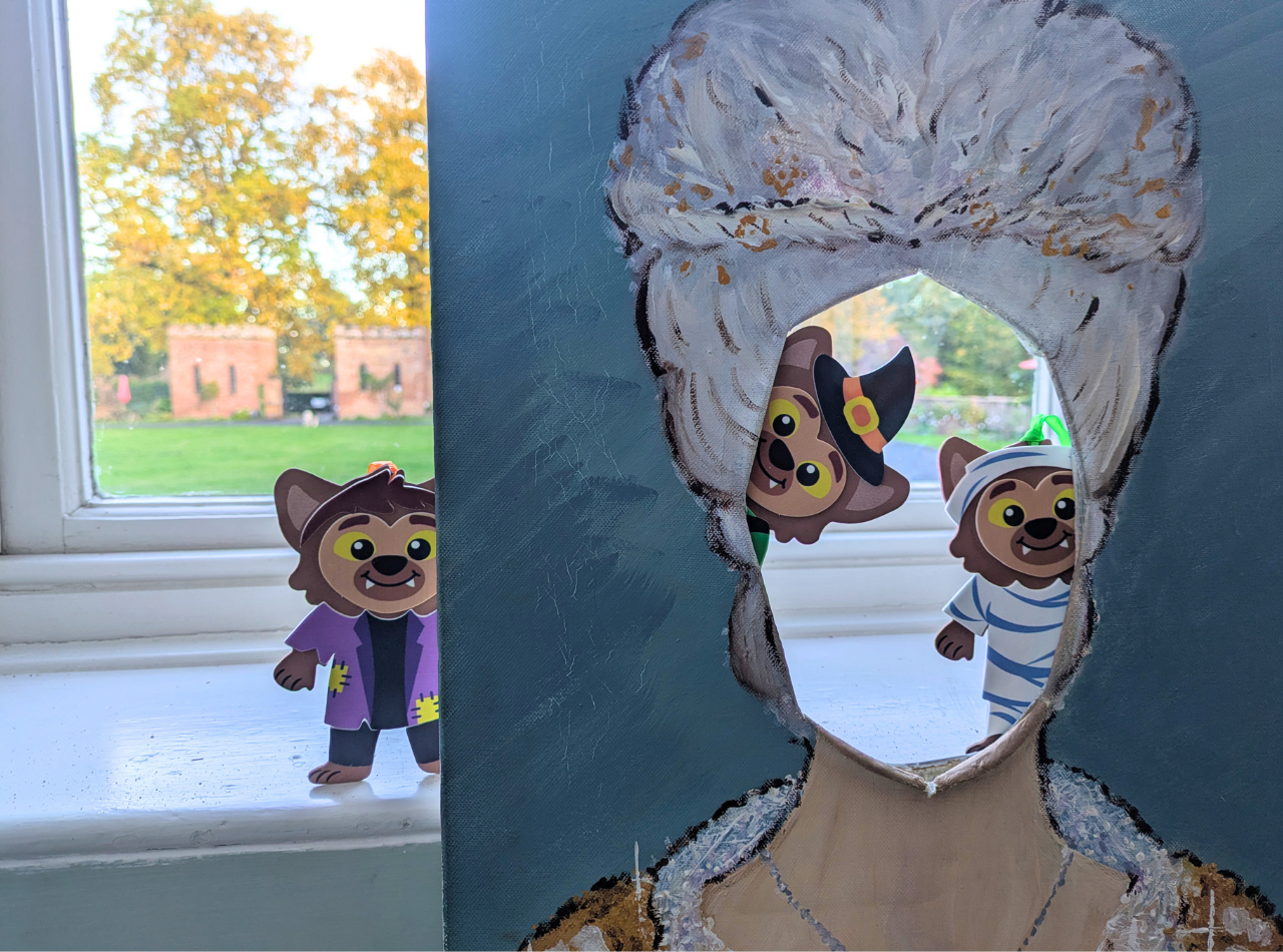 Miniature werewolf characters in Halloween costumes peering around a face-in-hole board of Queen Charlotte, with the Castle bothies seen through the window behind