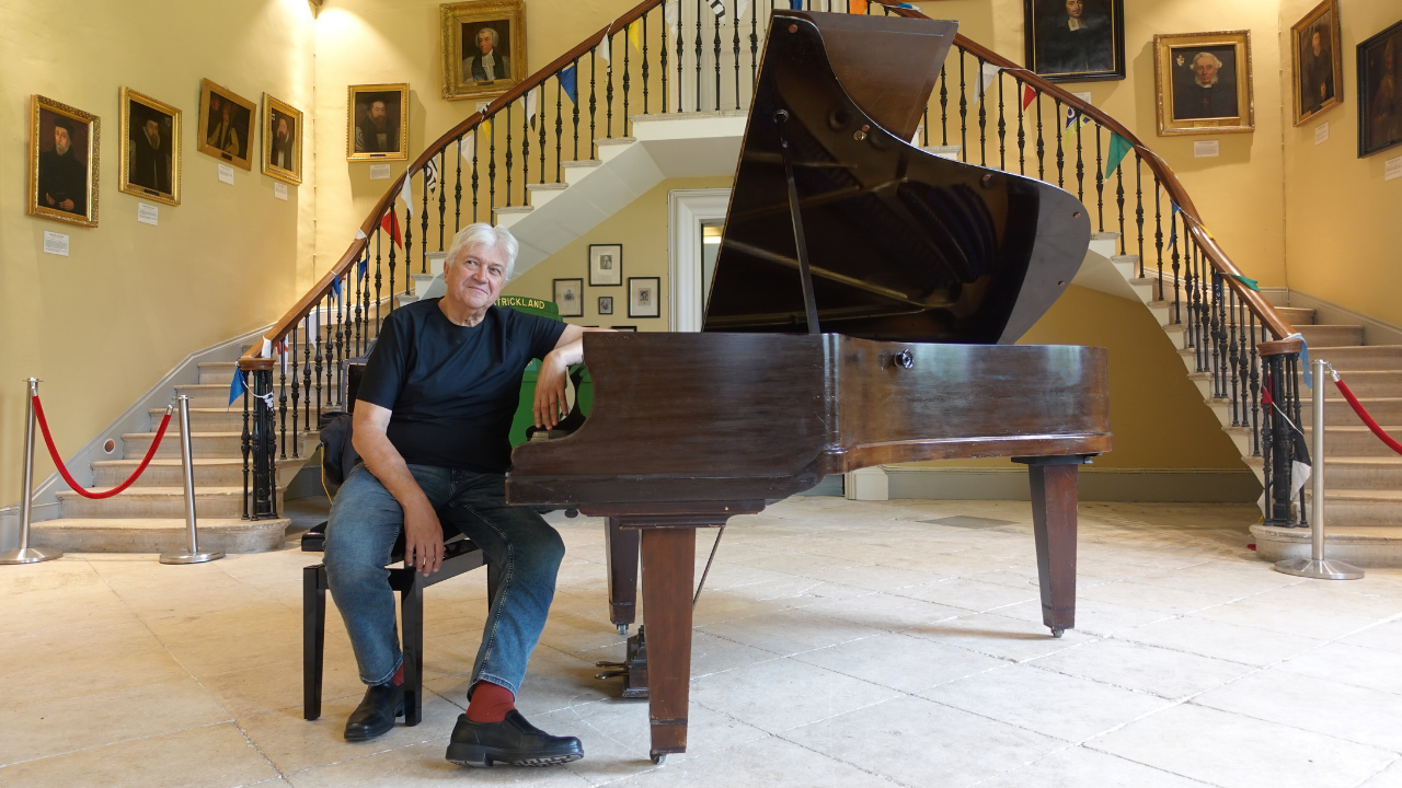 Older gentleman sitting at and leaning on a grand piano in the Castle's Great Hall