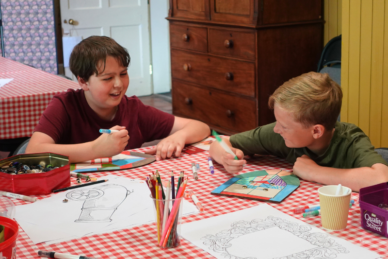 Two boys smiling at each other while colouring on wooden shields at a gingham craft table
