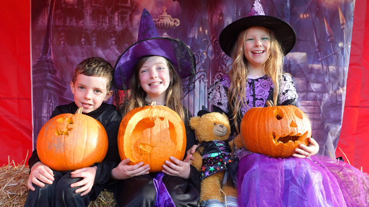 A boy and two girls sitting in front of a spooky backdrop, dressed for Halloween and holding carved pumpkins