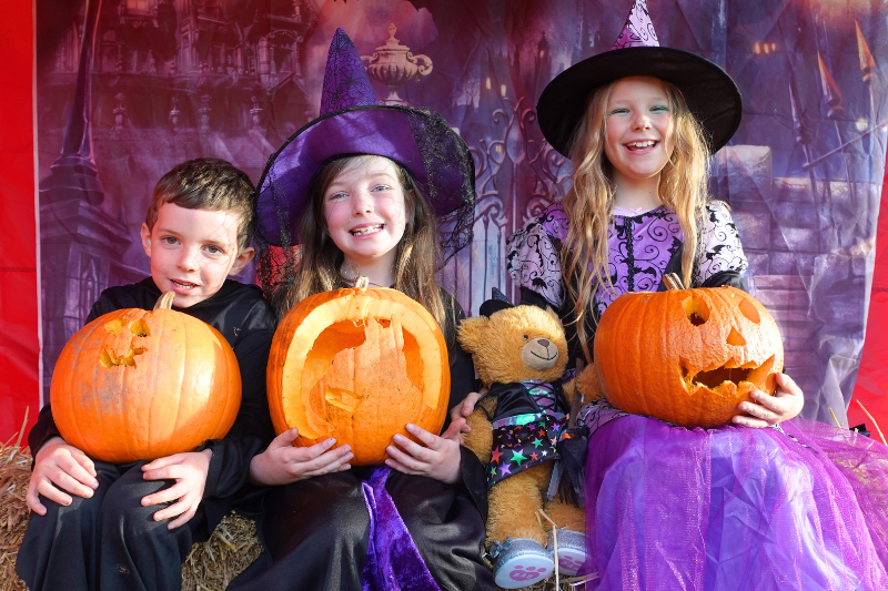 A boy and two girls sitting in front of a spooky backdrop, dressed for Halloween and holding carved pumpkins