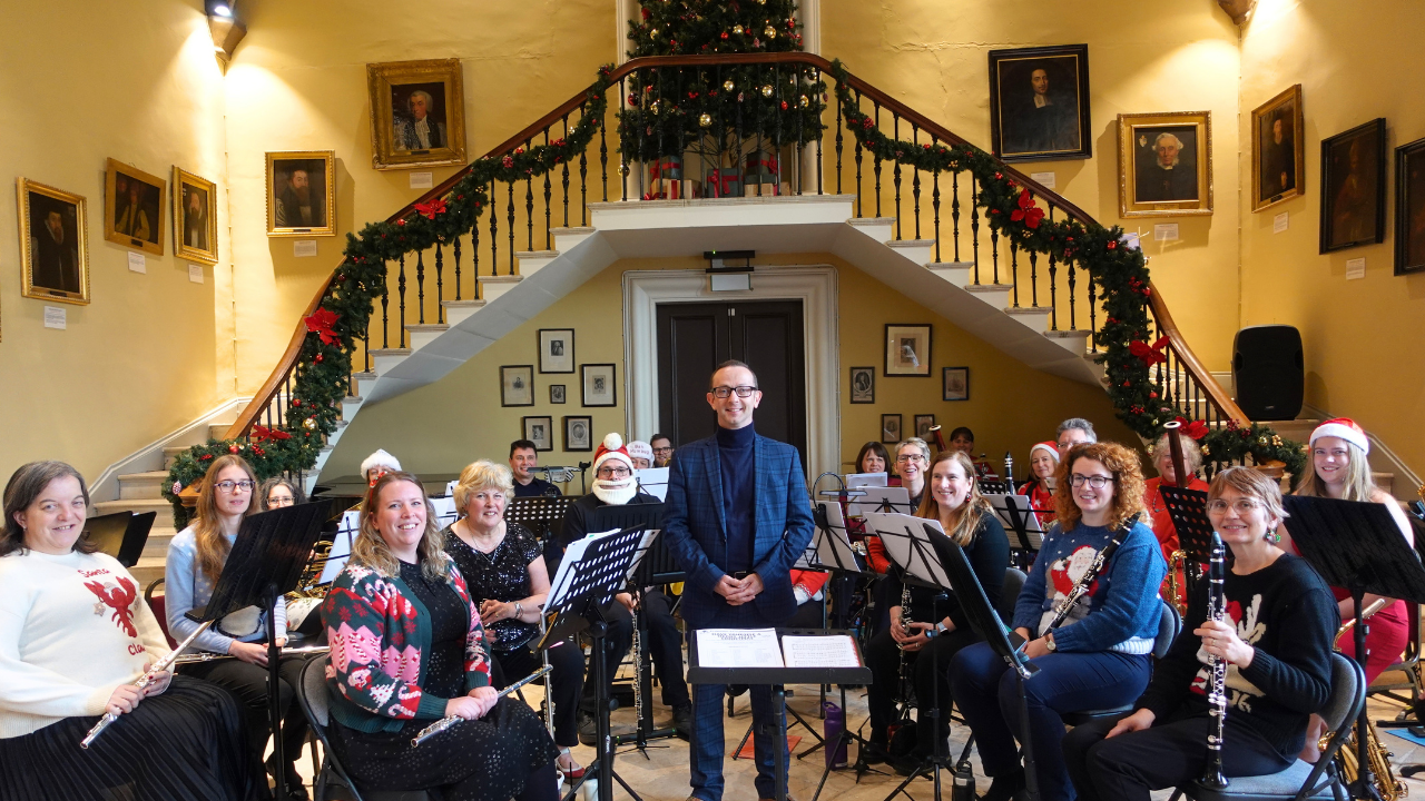 Worcestershire Winds Concert Band, smiling for a photo as they rehearse in the Castle's Great Hall with a Christmas tree and staircase garland behind them