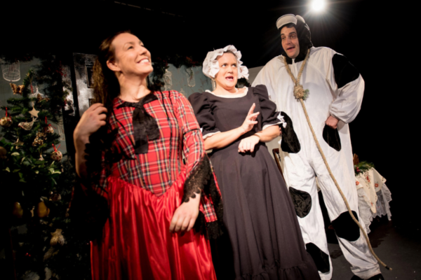 Three actors performing on a festive stage as a cow, servant and woman in a tartan print dress