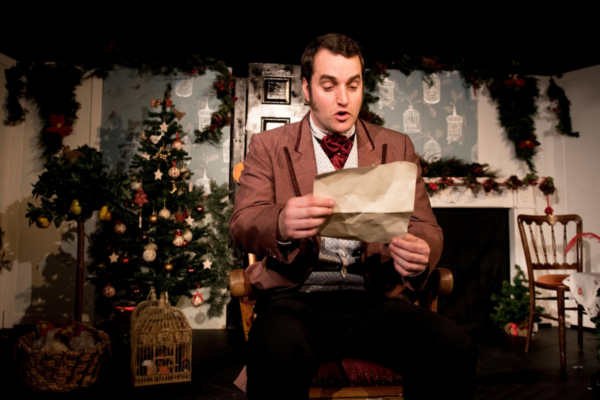 A male actor performing on a festive stage, sitting in a smart jacket and reading a letter aloud